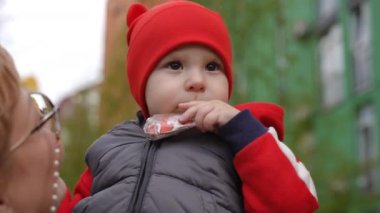 Ağır çekim. Yakın plan. Küçük bir çocuk elinde plastik bir oyuncak tutuyor ve dikkatlice uzaklığa bakıyor. Anne bir bahar günü dışarıda dikilirken bebeğini kollarına alır.