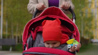 Ağır çekim. Kırmızı şapkalı küçük bir çocuğun bahar günü şehir parkında oturduğu bebek arabasıyla gezen bir anne. Anne durur ve çocuğun üzerine eğilir, gülümser ve konuşur.