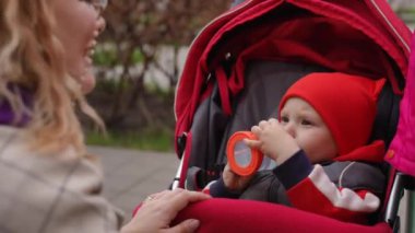 Ağır çekim. Yakın plan. Küçük bir çocuk dışarıdaki bir bebek arabasında otururken plastik bir şişeden su içerken, annesi ona sevgiyle bakıyor. Mutlu anne çocuğunu öper.