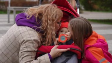 Ağır çekim. Anne ve kızı, dışarıda bebek arabasında otururken plastik şişeden su içen küçük kardeşiyle öpüşüyor. Küçük bir çocuk su içiyor, annesi ve kız kardeşi çömeliyor.