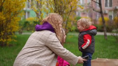 Ağır çekim. Küçük bir çocuk şehir parkındaki sarı çiçekli çalıların arasında oynarken annesi ve genç kızı çimenlerde oturup onu izliyorlar.