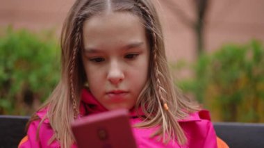 Ağır çekim. Yakın plan. Pembe spor ceketli bir kız öğrenci apartmanın yanındaki bankta oturuyor ve akıllı telefonun ekranına bakıyor.