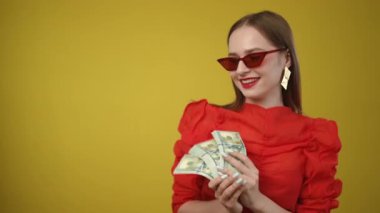 Kırmızı elbiseli ve güneş gözlüklü şık bir kadın, parlak sarı arka planda nakit gösteriyor.