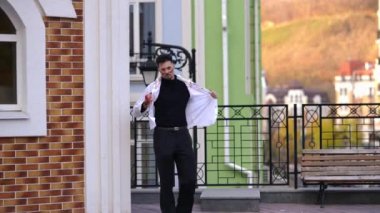 Beyaz gömlek giyen ve maske takan bir adam, çağdaş bir şehir binasının önünde telefonuyla meşgul olarak duruyor.