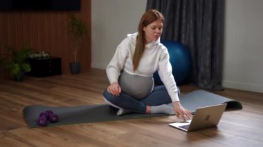 Huzurlu evinde hamile bir kadın hem meditasyon yapıyor hem de laptopunu kullanıyor.