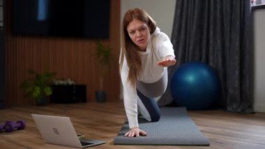 Evde odaklanmış bir kadın yoga yapıyor, özenle bilgisayarını kullanarak sanal egzersiz yapıyor.