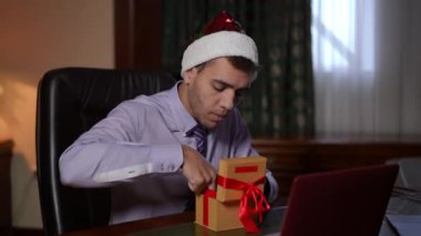 Noel Baba şapkalı bir adam masasında renkli bir hediyeyi neşeyle açıyor, tatil neşesini iş hayatıyla harmanlıyor.