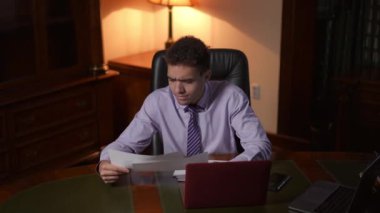 Resmi tişört giymiş bir adam sofistike bir ofis ortamında derinden ve dikkatli bir şekilde çeşitli evrakları analiz ediyor.