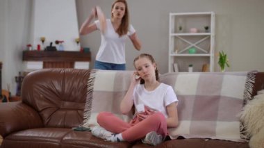 Bir anne, akıllı telefonuyla dikkati dağılmış bir şekilde kanepede oturan genç kızını azarlarken hayal kırıklığını ifade eder. Sahne, jenerasyon gerilimini ve iletişim boşluğunu...