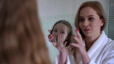 Beyaz bornozlu bir anne banyoda küçük bir ayna tutuyor ve yüzüne krem sürerken genç kızının yansımasına odaklanıyor. Samimi banyo ayarları bir sabahı aydınlatıyor.