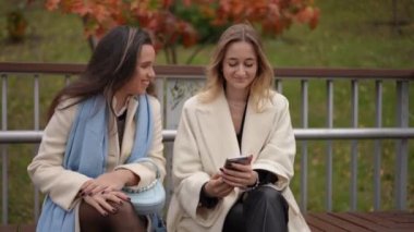 İki kadın, neşeli bir park ortamında telefonlarını kontrol ederken neşeli bir anı paylaşıyorlar.