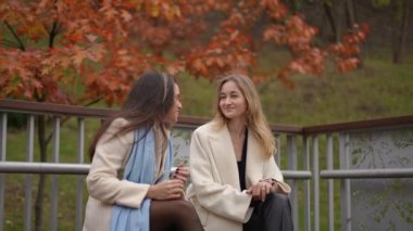 Şık paltolu iki neşeli kadın, güzel bir sonbahar ortamında canlı yapraklarla birlikte gülüyorlar.