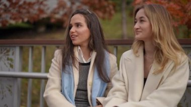 İki şık giyinmiş kadın, güzel, manzaralı bir sonbahar ortamında neşeli bir anı paylaşıyorlar.