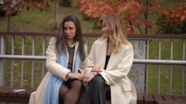 İki kadın, ahşap bir bankta oturmuş, açık havada canlı bir sonbahar yeşilliği arasında rahat bir anın tadını çıkarıyorlar.