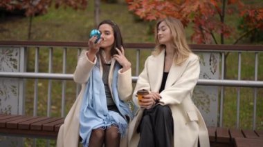 İki şık kadın arkalarına yaslanıp güzel bir bankta rahatlar, içkilerini yudumlar ve canlı sonbahar yapraklarının tadını çıkarırlar.