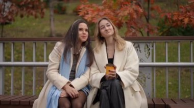 İki şık giyimli kadın sevilen bir bankta oturmuş renkli sonbahar yaprakları arasında canlı hikayeler anlatıyorlar.