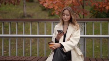 Bir kadın, sakin bir sonbahar parkında tesadüfen telefonunu kullanırken sıcak bir fincan kahveden zevk alır.
