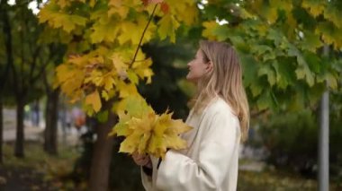 Neşeli genç bir kadın güzel bir sonbahar ortamında canlı ve renkli yaprakları tutarken geniş bir gülümseme sergiliyor.