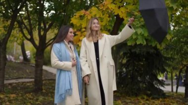 İki kadın parktaki renkli sonbahar yaprakları altında sohbet ediyor, hem moda hem de mevsimsel güzellikleri sergiliyorlar.