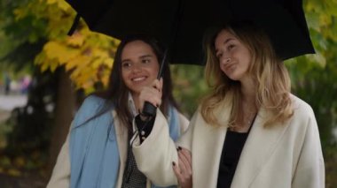 İki genç kadın, canlı sonbahar yaprakları arasında birlikte yürürken bir şemsiyeyi paylaşıyorlar.