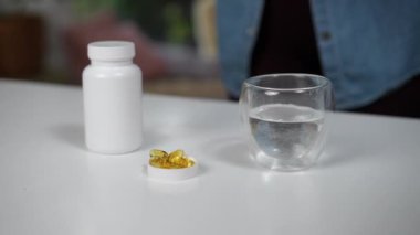 Bir el, yanında bir bardak su olan bir şişeden altın renkli besin takviyeleri seçiyor. Bu görüntü, sağlıklı bir günün parçası olarak takviye madde alma eylemini vurgular.