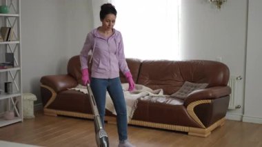 Sıradan giyinen, oturma odasını süpüren, evlerin temizliğini ve düzenini gururla sergileyen bir kadın.