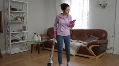Pembe eldivenli bir kadın telefonunu kontrol ederken oturma odasını süpürüyor. Temizliği çoklu görevle harmanlıyor..