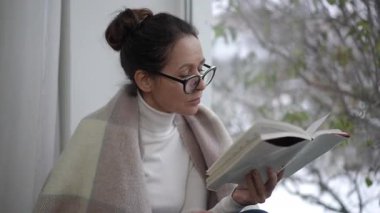 Yumuşak bir battaniyeye sarılmış bir kadın güneşli pencerenin yanında kitap okurken huzurlu bir anda zevk alır.