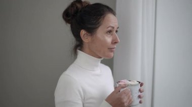 Rahat bir kazak giyen bir kadın sıcak kakaosunu yudumlarken düşünceli bir şekilde bir kış günü pencereden dışarı bakar.