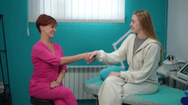 Pembe üniformalı gülümseyen kozmetik uzmanı klinikteki genç bayan müşteriyle el sıkışıyor. Muayeneden önce modern güzellik tedavi odasında dostane bir atmosfer..