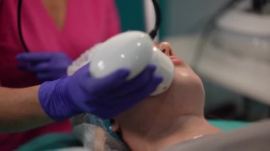 Estetisyen, yüz tedavisi sırasında genç kadınların yüzünde gelişmiş bir cihaz kullanıyor. Teknisyen cilt teknolojisi uygularken müşteri gözleri kapalı yatıyor..