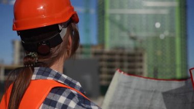 Çalışkan bir inşaat işçisi güneşli gökyüzünün altındaki bir inşaat alanını denetlerken planlarını da inceler.