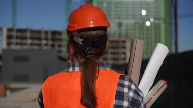 Güvenlik yeleği ve miğfer takmış bir inşaat işçisi elinde ayrıntılı planlar tutarken telefonda konuşuyor.