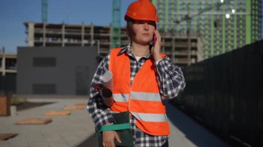 Güvenlik kaskı ve parlak turuncu yelek giyen bir inşaat işçisi, hareketli bir inşaat alanında akıllı telefon kullanır.