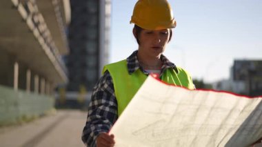 Bir inşaat işçisi parlak güneş ışığı altında işlek bir şantiyede mimari planları dikkatle inceler.