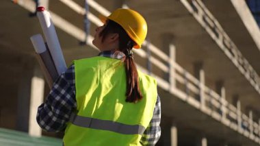 Yoğun bir şantiyede, güvenlik donanımı giyen bir inşaat işçisi ayrıntılı planları dikkatle gözden geçirir.