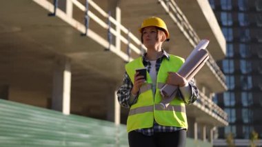 Güvenlik donanımı giyen bir inşaat işçisi, yoğun ve aktif bir inşaat sahasında ayrıntılı planları dikkatle gözden geçirir.