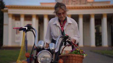 Tarz sahibi bir kadın güzel motosikletiyle neşe içinde etkileşime girer, güzel bir çiçek sepetiyle süslenir.