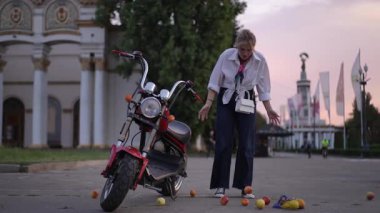 Bir kadın mutlu bir şekilde mobiletinin yanında düşen elmaları topluyor ve muhteşem bir günbatımına karşı duruyor.