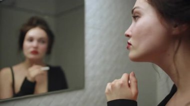 Siyah saçlı genç bir kadın banyodaki aynadaki yansımasına bakarken dudaklarına sigara tutuyordu. Görüntü onun ön plandaki profiline odaklanır, alevi bulanık arkaplanda görünür hale getirir.