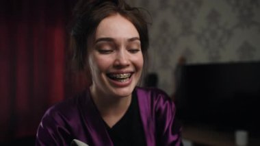 Mor saten bir bornozla, diş telleri kontrolsüz kahkahalara boğulmuş, gözleri yarı kapalı genç bir kadın. Sahne duygusal çöküntünün kırılgan anını yakalar, gözyaşları ve zorla neşe arasında sallanır..