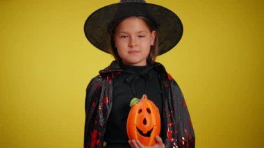 Şirin giyinmiş genç bir cadı parlak turuncu bir balkabağı tutuyor, Cadılar Bayramı ruhunu ve neşeli hareketlerini gösteriyor.