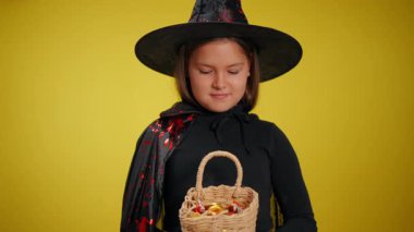 Kostümlü genç bir cadı elinde bir sepet şeker tutarken gülümsüyor, canlı sarı bir zemin üzerine kurulmuş..