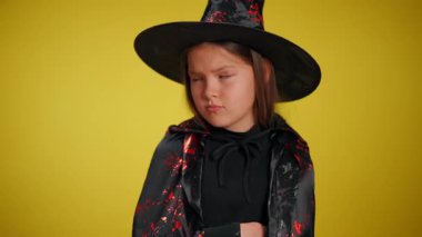 Cadı kostümlü, siyah pelerinli ve şapkalı neşeli bir kız parlak sarı bir arka planda duruyor.