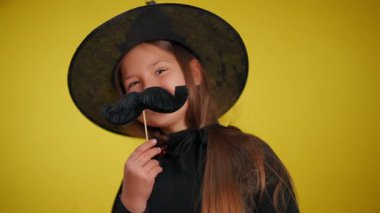 Cadı gibi giyinmiş genç bir kız neşeli ruhunu canlı sarı bir arka plana karşı eğlenceli aksesuarlarla sergiliyor..