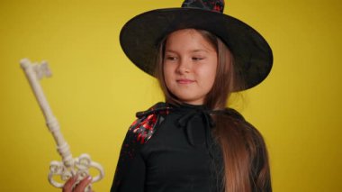 Genç bir kız, kaprisli bir cadı gibi giyinmiş, büyük, parlak bir anahtar tutuyor, bir eğlence ve sihir teması sergiliyor.