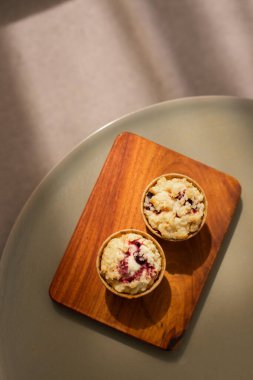 İki yeni pişmiş kek, ahşap bir tahtanın üzerinde, parçalanmış tepeleri ve böğürtlen parçalarıyla, yumuşak güneş ışığıyla yıkanmış, rahat, nötr bir zemin üzerine kurulmuş..