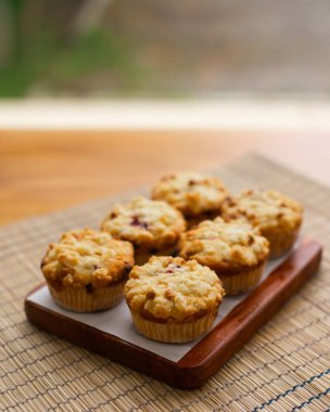 Six freshly baked muffins sit on a wooden tray lined with parchment paper. They have a crumbly, golden-brown topping with hints of fruit, possibly berries. The woven mat background gives the image a cozy, homemade feel. clipart