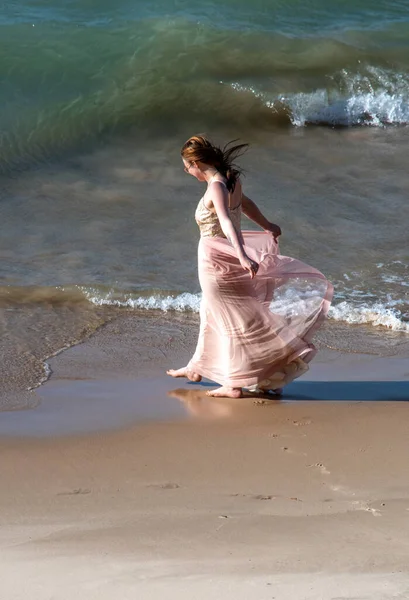 Barefoot young woman in a flowing dress, walks along the lake as waves rush the beach