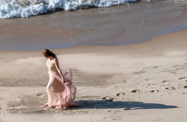 Young woman in a flowing dress walks along the beach as waves rush in